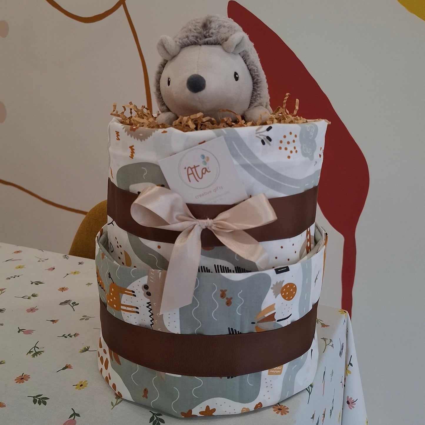 Three-tiered diaper cake with a plush hedgehog toy on a floral tablecloth.