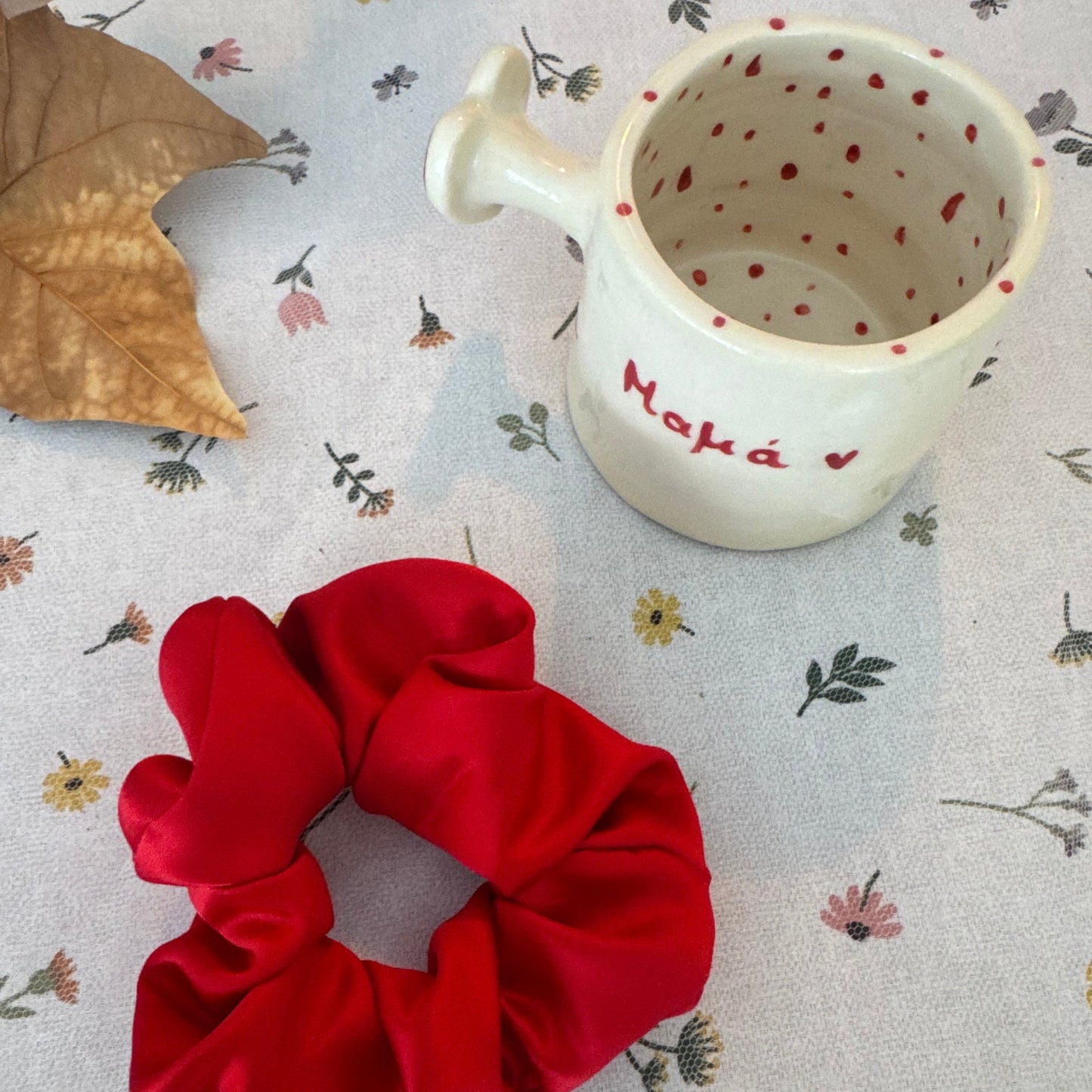 Red scrunchie on a patterned surface with a small cup saying @mum@ in greek and leaf.