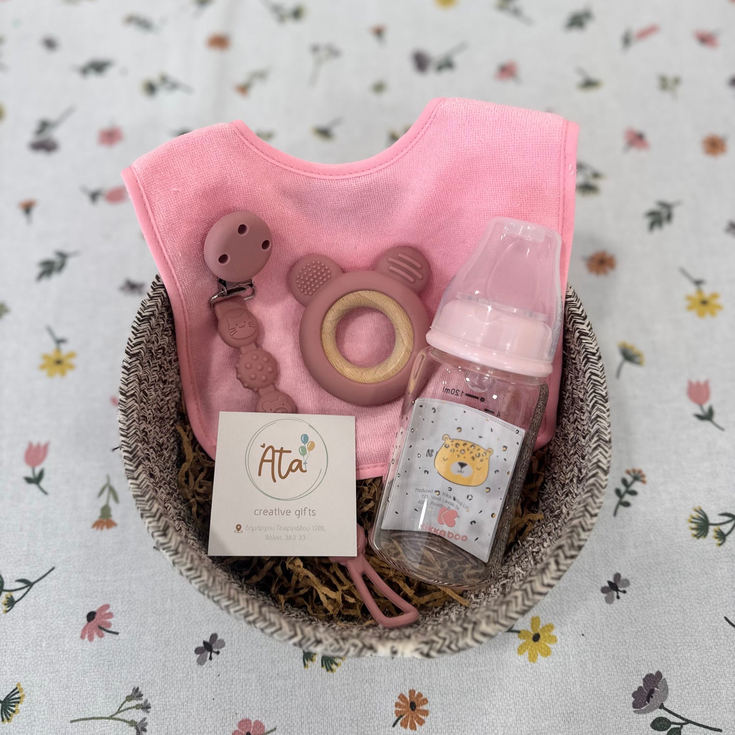 Baby gift set with pink bib, bottle, and card in a basket on a floral background