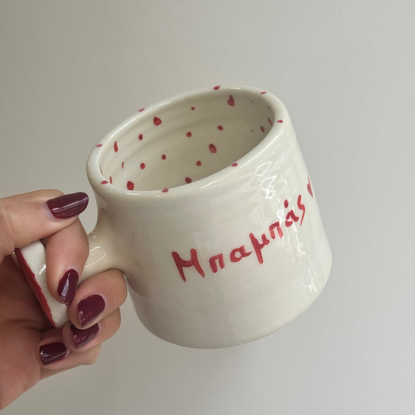 White mug with red polka dots and text held by a hand against a plain background