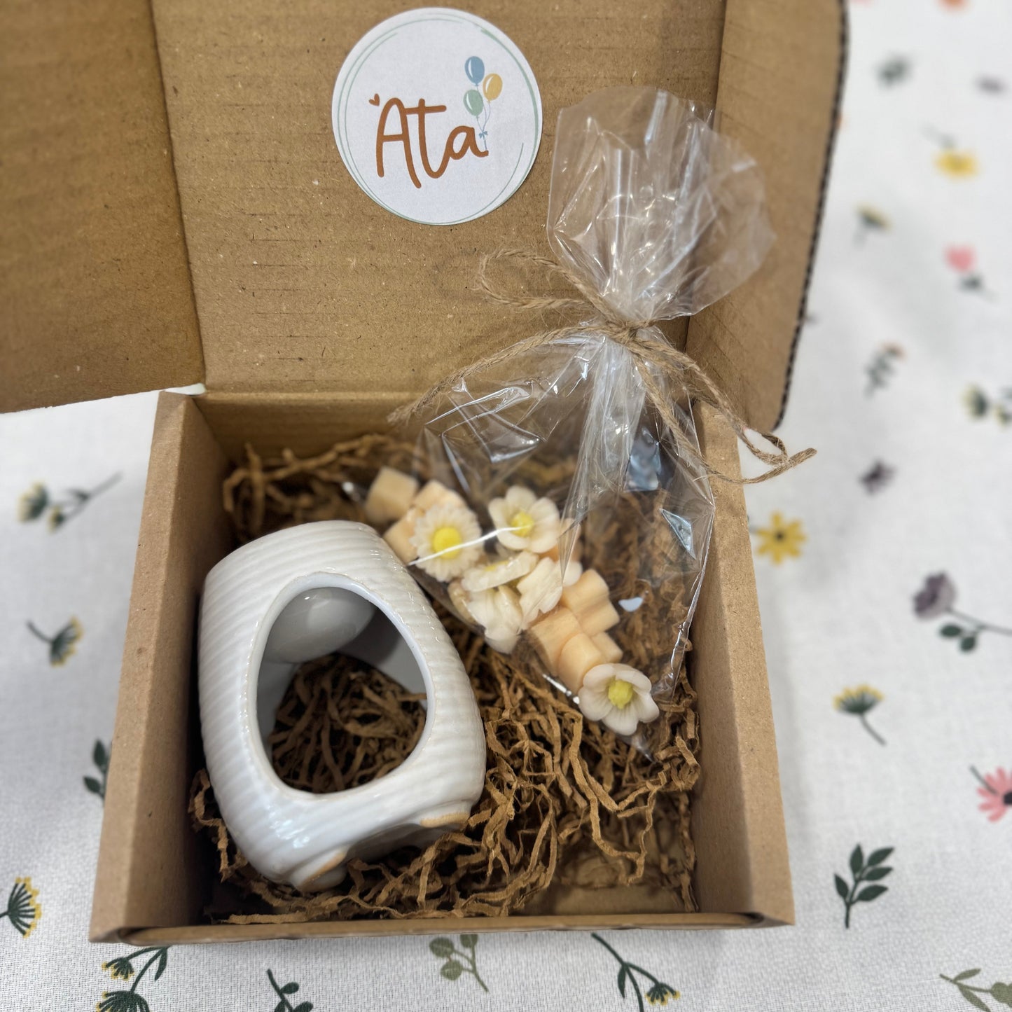 Gift box with a white ceramic item, flowers shaped wax melts, and decorative straw on a floral-patterned surface.