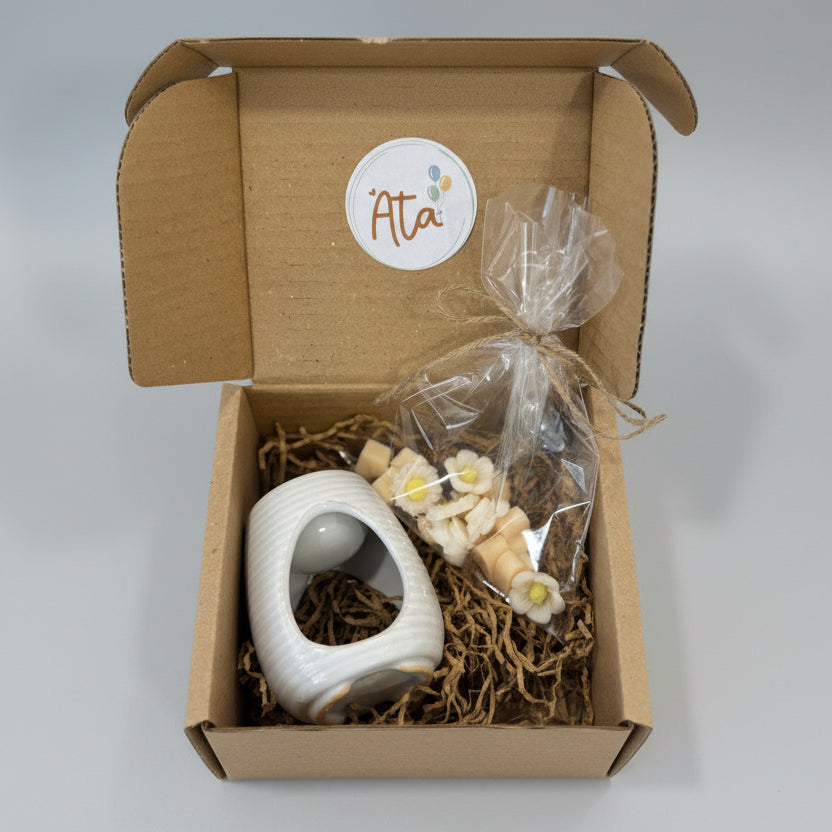 Gift box with a white ceramic item, flowers shaped wax melts, and decorative straw on a floral-patterned surface.