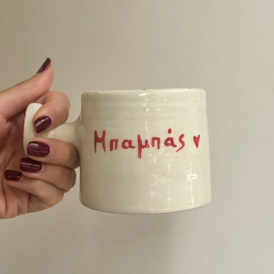 Hand holding a white mug with red text "dad" in greek against a plain background