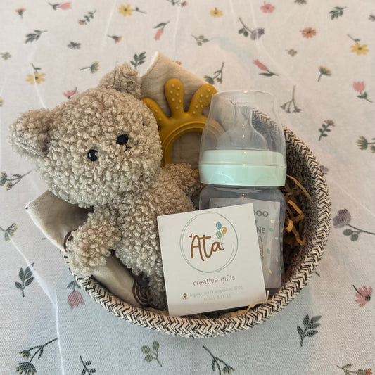 Plush toy grey bear with a teething ring and a bottle on a floral blanket, branded 'Ata'.