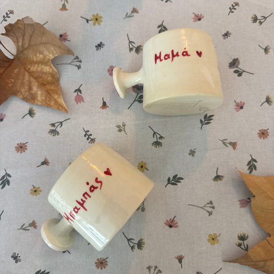 Two ceramic cups with text mum and dud in greek on a patterned fabric background