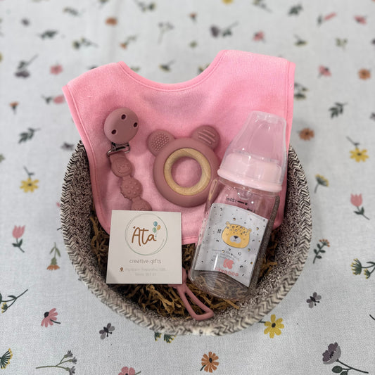 Baby gift set with pink bib, bottle, and card in a basket on a floral background