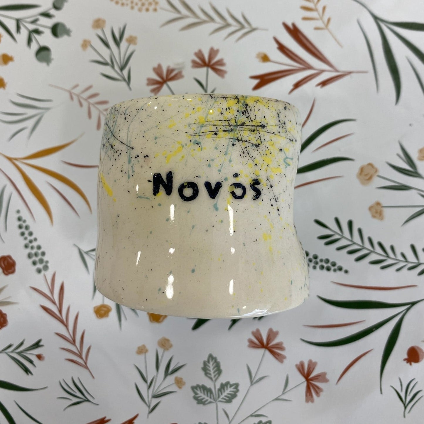 Ceramic container with 'Novos' saying on a floral-patterned background