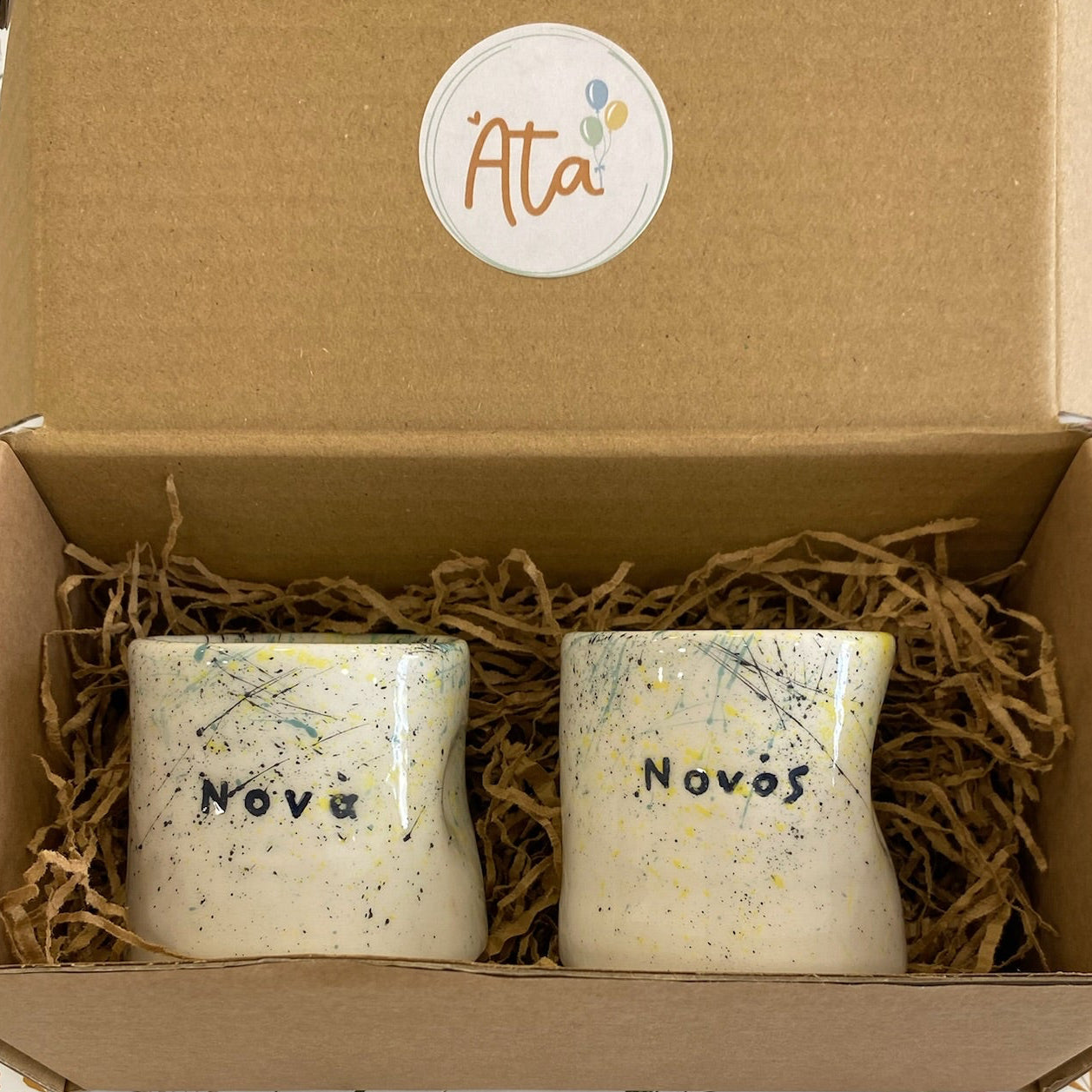 Two small containers labeled 'νονός' and “Νονά “in a brown box with 'Ata' branding on a floral-patterned surface.