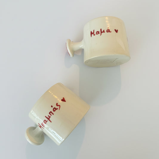 Two ceramic cups with text mum and dud in greek on a patterned fabric background