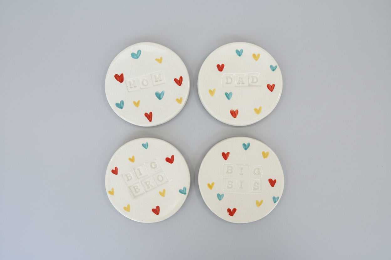 White ceramic coasters with 'DAD', 'MOM', 'BIG BROTHER', and 'BIG SISTER' text on grey background 