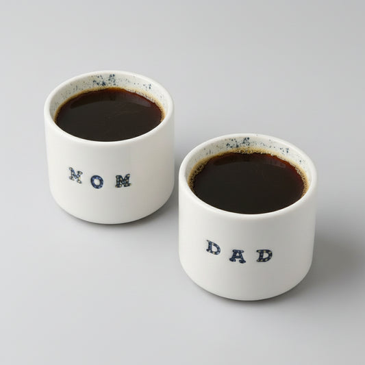 Two white ceramic pots with 'MOM' and 'DAD' engraved on them, placed on a grey background illed with coffee